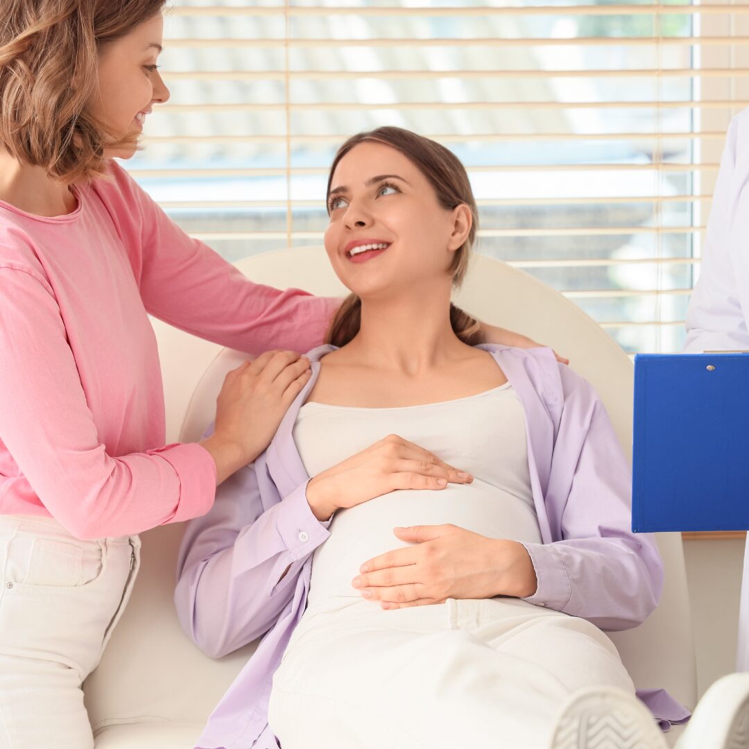 A same-sex couple one showing support to the other lying on a chair pregnant.